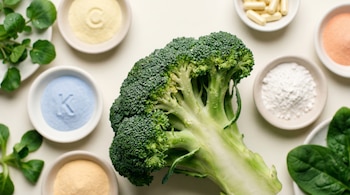 Una cabeza de brócoli verde fresco con gotas de agua, rodeada por pequeños platos de cerámica con polvos de colores, cápsulas y hojas de berro y espinaca.