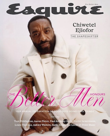 Chiwetel Ejiofor viste un abrigo blanco con botones oscuros y posa sentado sobre una silla de madera frente a una pared clara