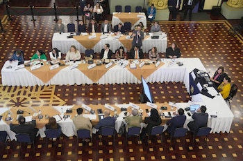 Personas sentadas en mesas dispuestas en forma de U con documentos y micrófonos en un salón con suelo de mosaico