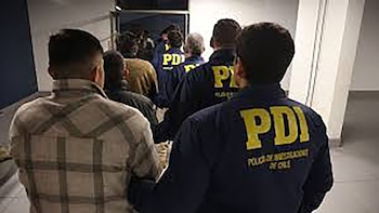 Vista trasera de varias personas, incluyendo agentes de la PDI con chaquetas azules, caminando por un pasillo blanco en una fila