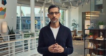Sundar Pichai, con gafas y cárdigan oscuro, de pie frente a ventanas grandes en un edificio moderno. El logo de Google es visible fuera