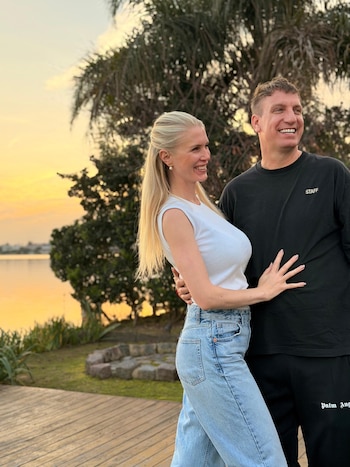 Una mujer rubia con camiseta blanca y jeans abraza a un hombre con camiseta negra al aire libre junto al agua durante el atardecer