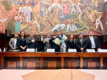 Diez personas posan detrás de una mesa de madera; el hombre central sostiene dos cinturones de boxeo verdes del CMB. Detrás hay un mural colorido