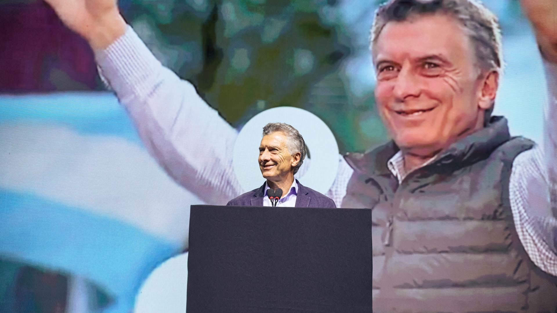 El expresidente argentino Mauricio Macri sonríe desde el podio, con una imagen de sí mismo levantando los brazos proyectada de fondo, durante un acto de comunicación.