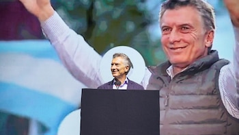 Mauricio Macri sonriendo detrás de un atril negro, con una gran pantalla de fondo que muestra su imagen con los brazos levantados