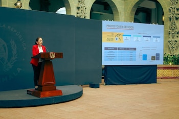 Mujer con saco rojo y anteojos de pie frente a un atril de madera. Detrás, una pantalla grande muestra gráficos y datos sobre proyectos