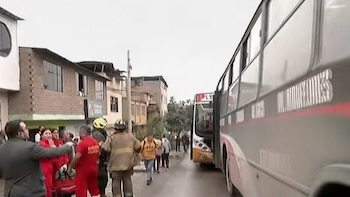 Dos autobuses de transporte público chocados en una calle mojada, con personal de bomberos y de emergencia en uniformes rojos y personas civiles alrededor