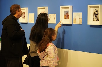 Un hombre, una mujer y una niña observan cuadros enmarcados en una pared azul y blanca. La niña señala una de las obras exhibidas