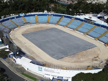 Vista aérea de un estadio con gradas azules y amarillas, una pista de atletismo de tierra, un campo central cubierto de gris y árboles