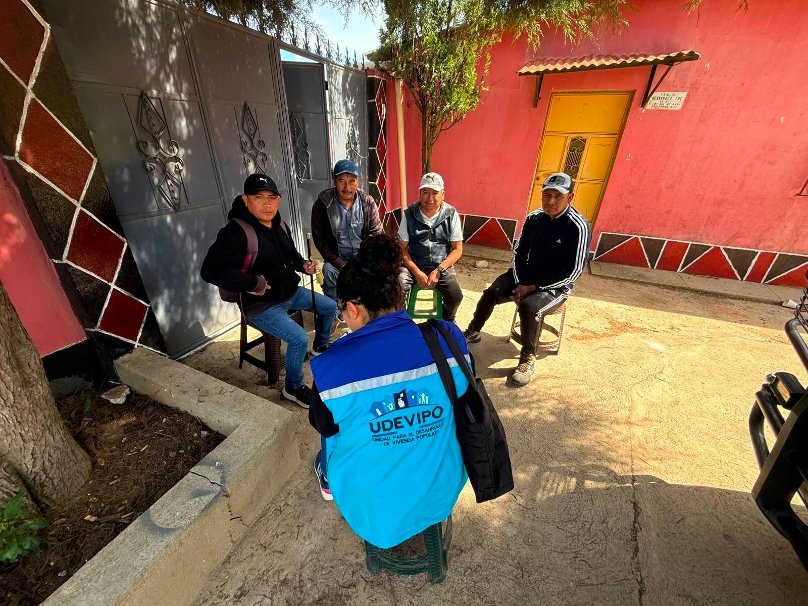 Una representante de la Unidad para el Desarrollo de Vivienda Popular (UDEVIPO) dialoga con cuatro hombres sentados en un patio. (Ministerio de Comunicaciones, Infraestructura y Vivienda)