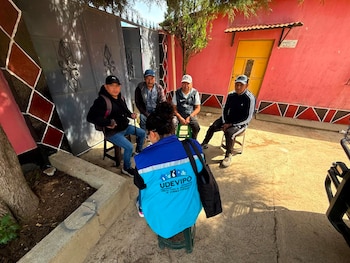 Cinco personas sentadas al aire libre: una mujer con chaleco UDEVIPO de espaldas, cuatro hombres al frente. Detrás hay una pared rosa y un portón metálico gris