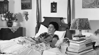 Fotografía en blanco y negro de Frida Kahlo recostada en una cama de cuatro columnas junto a una mesa con libros y una lámpara