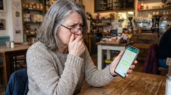 Mujer de mediana edad con gafas y cabello gris sentada en una cafetería. Sostiene un smartphone con WhatsApp y su mano cubre parcialmente su boca.