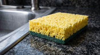 Esponja porosa amarilla con fibra verde sobre encimera de granito oscuro junto a un fregadero de acero inoxidable.