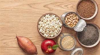 Una batata, una manzana roja, un cuenco con palomitas, legumbres enlatadas, semillas de chía, lentejas y avena sobre madera.