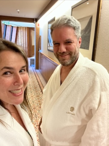 Malena Guinzburg y Adrián Montesoro en batas blancas. Fondo de pasillo con pisos estampados, zócalos de madera y cuadros en la pared