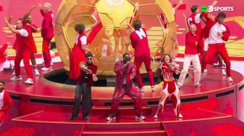 Artistas en trajes rojos y blancos bailando en un escenario rojo. Un cantante central viste un traje estampado. Al fondo, una gran esfera dorada con forma de balón de fútbol