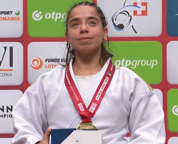 Una mujer vestida con judogi blanco sostiene un premio pequeño y lleva una medalla al cuello frente a un panel con logotipos