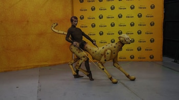 Un intérprete porta una estructura mecánica y vestuario de guepardo frente a un fondo amarillo con logotipos del musical El Rey León