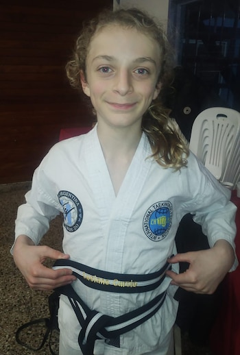 Retrato de un joven sonriente, de tez clara y cabello rizado, con un dobok blanco de Taekwondo y ajustando su cinturón negro bordado