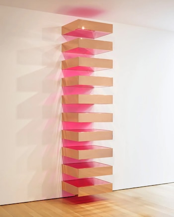 Una escultura minimalista vertical con diez cajas rectangulares de color cobre y rosa vibrante, montada en una pared blanca y suelo de madera