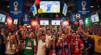 Multitud de personas animando y levantando vasos de cerveza en un bar. Hay pantallas mostrando un partido de fútbol, banderas de la Copa Mundial FIFA 2026, confeti y globos.
