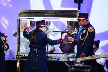 Dos jóvenes con uniformes de gala azul marino y dorado participan en el Sorteo Magno de la Lotería Nacional, transfiriendo un palillo con un número