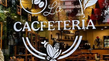 Vista exterior de la CACfetería con el nombre y logo blanco en el cristal, revelando mesas de madera, sillas y estantes con productos en el interior