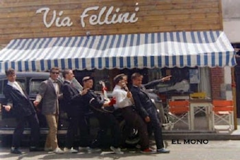 Seis hombres con traje posan alineados frente a un comercio con toldo a rayas y la inscripción Vía Fellini