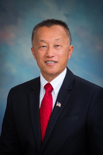 Retrato de un hombre asiático de mediana edad con traje oscuro, camisa blanca, corbata roja y pin de bandera de EE. UU. en un fondo azul degradado