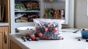 Una bolsa transparente con fresas, arándanos y moras congeladas sobre una encimera blanca, junto a un congelador abierto.