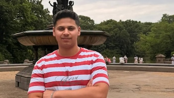 Hombre joven con camiseta de rayas rojas y blancas y pulsera plateada. Detrás hay una fuente de piedra, árboles verdes y personas borrosas junto a un estanque