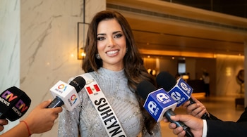 Suheyn Cipriani sorri ao entrevistar vários microfones de imprensa. Ele veste um terno cinza brilhante e uma faixa branca com a bandeira do Peru e seu nome.