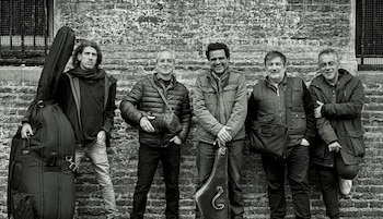 Cinco hombres de pie frente a una pared de ladrillos en blanco y negro. Dos de ellos sostienen fundas de instrumentos musicales