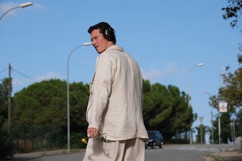 Un hombre con traje beige, auriculares negros y una rosa amarilla camina sobre una calle asfaltada, con árboles, farolas y cielo azul de fondo