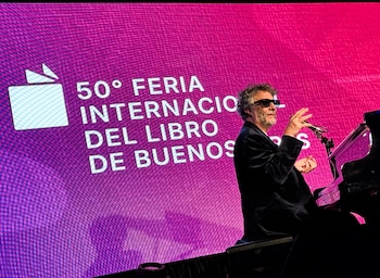 Hombre de cabello rizado y barba, con gafas de sol y traje oscuro, tocando el piano en un escenario con una pantalla gigante magenta detrás