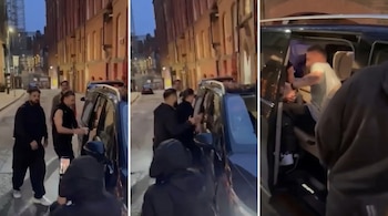 Captura de una persona con tatuajes entrando a una SUV oscura, rodeada de hombres, en una calle urbana con edificios de ladrillo iluminados por la noche.