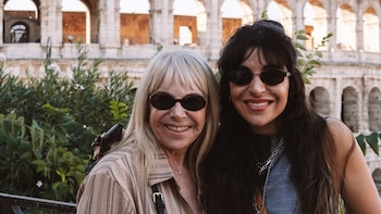 Dos mujeres sonrientes con gafas de sol posan en primer plano, con cabello claro a la izquierda y oscuro a la derecha, frente a la estructura del Coliseo de Roma