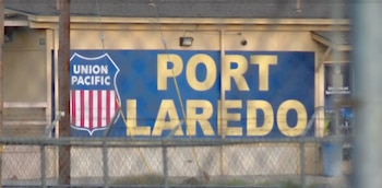 Un cartel azul con el logo de Union Pacific y la inscripción "PORT LAREDO" en letras amarillas, frente a un edificio y detrás de una valla