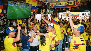Multitud de hinchas colombianos, vistiendo camisetas amarillas, celebran con entusiasmo en un bar decorado con banderas, viendo un partido de fútbol en TV.