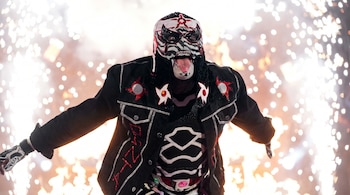 Hombre enmascarado con máscara decorada en blanco, negro y rojo, chaqueta negra con detalles plateados y rojos, guantes y lengua fuera; fondo de chispas brillantes.