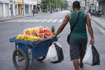 Hombre de piel morena de espaldas empuja un carro azul con plátanos, sandía y piña. Viste camiseta verde y pantalones cortos a cuadros, en una calle urbana.