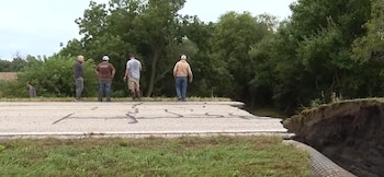 Cuatro hombres observan el borde cortado de una carretera de asfalto agrietada junto a un gran socavón y vegetación