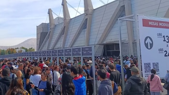Gran multitud de personas haciendo fila frente a un edificio moderno con estructuras de concreto al aire libre. Hay señales de "MESAS" y números. Montañas al fondo