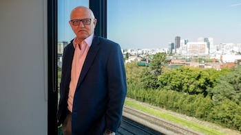 Hombre con gafas, camisa clara y saco azul de pie junto a un ventanal. Al fondo se observan vías de tren, árboles y edificios