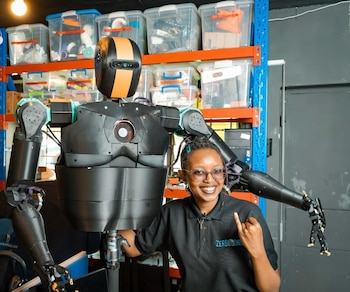 Una mujer sonriente con gafas y camiseta negra está junto a un robot humanoide de color negro y naranja en un taller