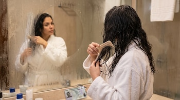 Mujer en albornoz blanco cepilla su cabello oscuro y mojado frente a un espejo de baño empañado. Una cédula de residencia colombiana se ve en el lavamanos.