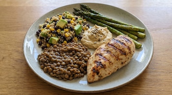 Un plato de comida con pollo a la parrilla, lentejas, ensalada de quinoa con maíz, frijoles negros y aguacate, hummus y espárragos verdes.