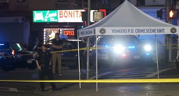 Tres agentes de policía y dos vehículos patrulla con luces intermitentes en una calle por la noche, con cinta de seguridad amarilla y carpa de unidad forense