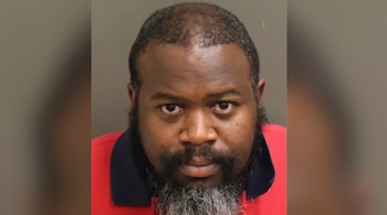 Fotografía policial de un hombre afroamericano de mediana edad, con barba y bigote canosos, y calvicie parcial, usando una camisa polo roja